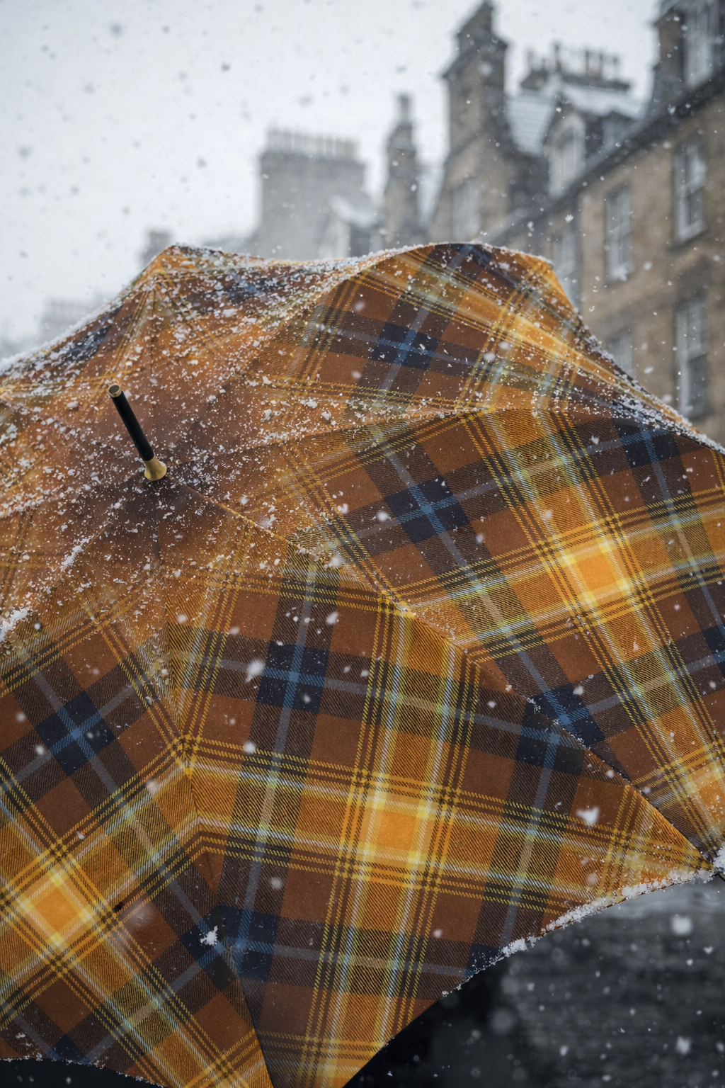 Close-up of tartan umbrella dusted with falling snow against historic stone buildings.