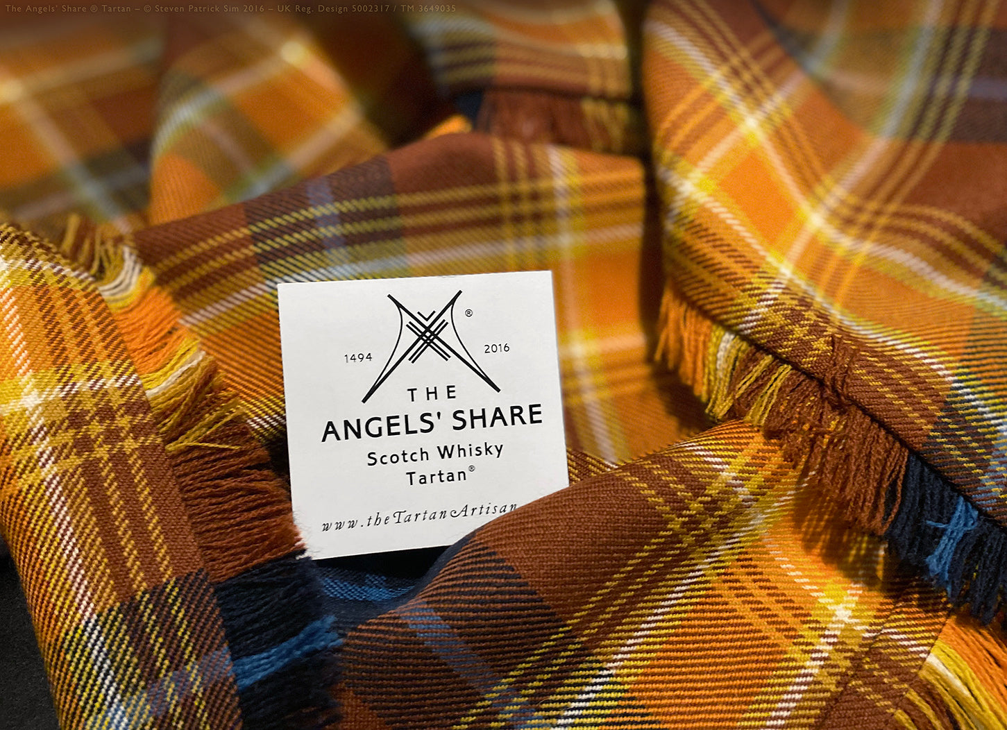 Fringed fly plaid in the Angels’ Share tartan with edition card nestled in folds. The Angels’ Share® fly plaid is shown styled with its official edition label — presented within the folds of the fabric, emphasising the authenticity and colour vibrancy.