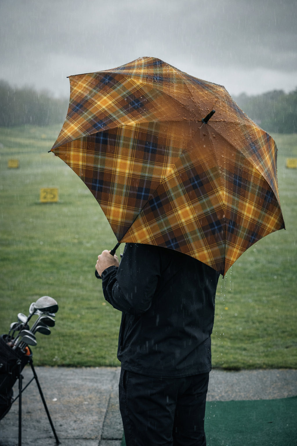 Rear three-quarter view of golfer holding the Angel’s Share tartan umbrella beside a golf bag, rain streaming off the rich amber and blue plaid canopy.