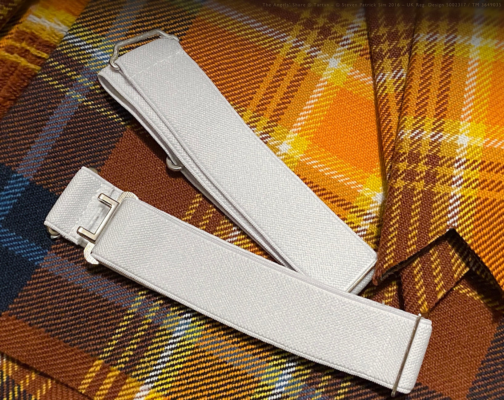 White kilt garters resting on the pleated fabric of the Angels' Share® tartan kilt. A clean pair of white formal kilt garters lies atop the vibrant pleats of the Angels' Share® Scotch Whisky Tartan. The luminous ochre, amber, and russet tones of the tartan highlight the garters' simplicity and contrast beautifully with the fabric’s rich colour story.