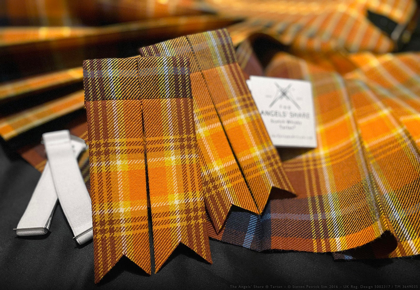 Matching tartan flashes and white elasticated garters displayed with a kilt made in the Angels’ Share® tartan. This image showcases a matching set of Angels’ Share® tartan flashes and pristine white garters — a complete accessory pairing to accompany a full kilt ensemble in this rich whisky-inspired design.