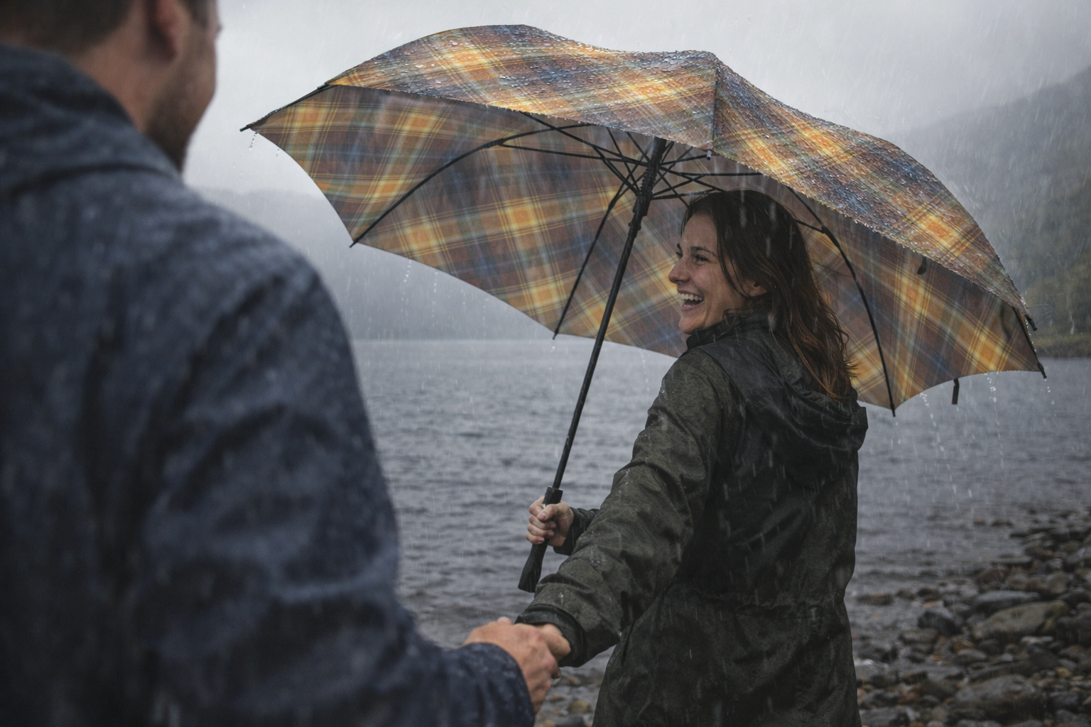Woman smiling under tartan umbrella by a loch in rain while holding partner’s hand.