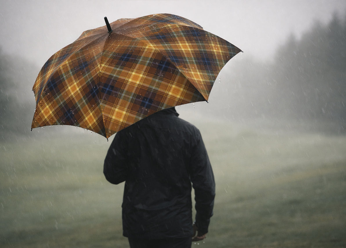 Rear view of golfer walking into misty rainfall beneath the Angel’s Share tartan umbrella, the bold amber plaid standing out against a muted green landscape.