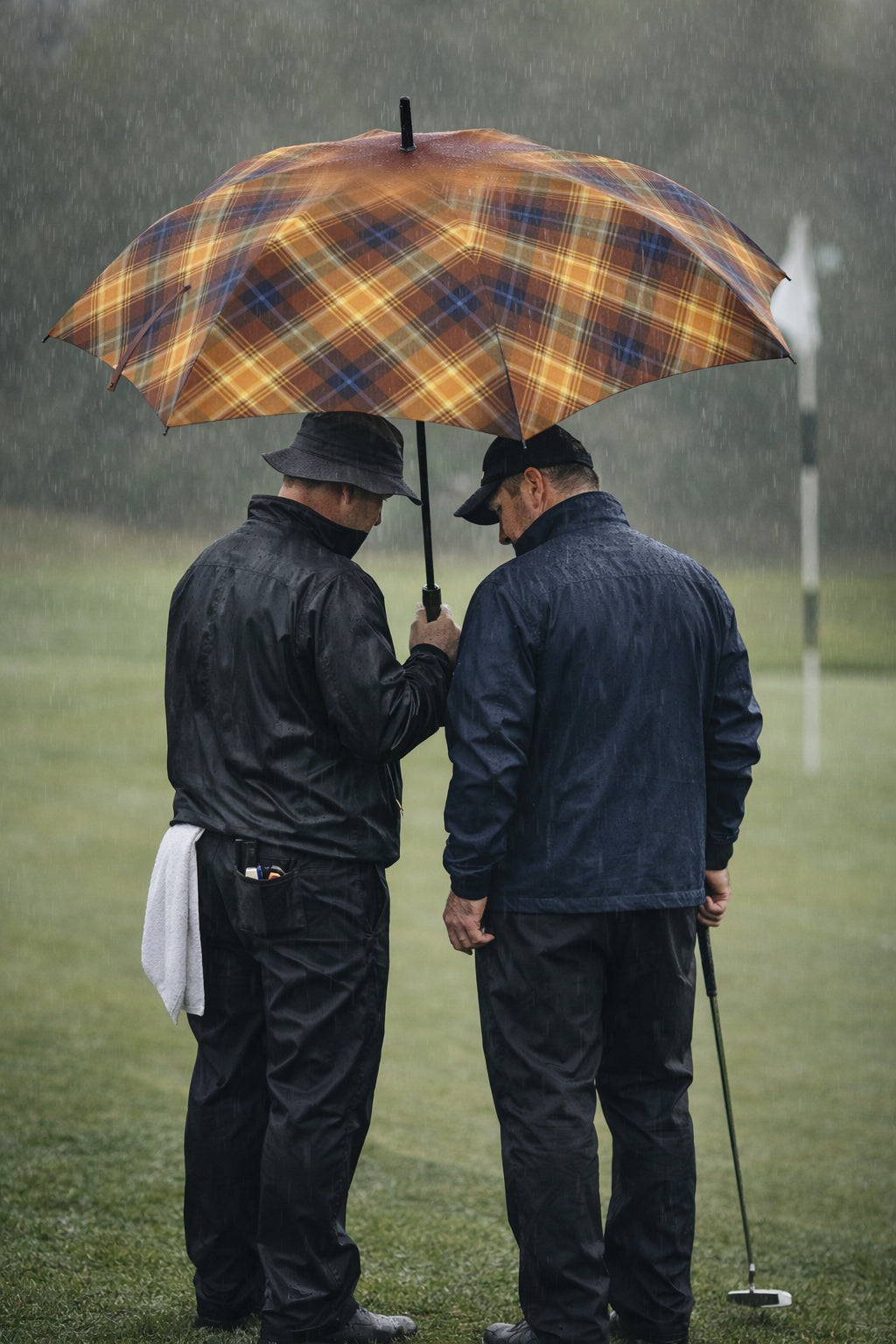Two golfers standing together under the wide Angel’s Share tartan umbrella during heavy rain, demonstrating generous canopy coverage on the course.