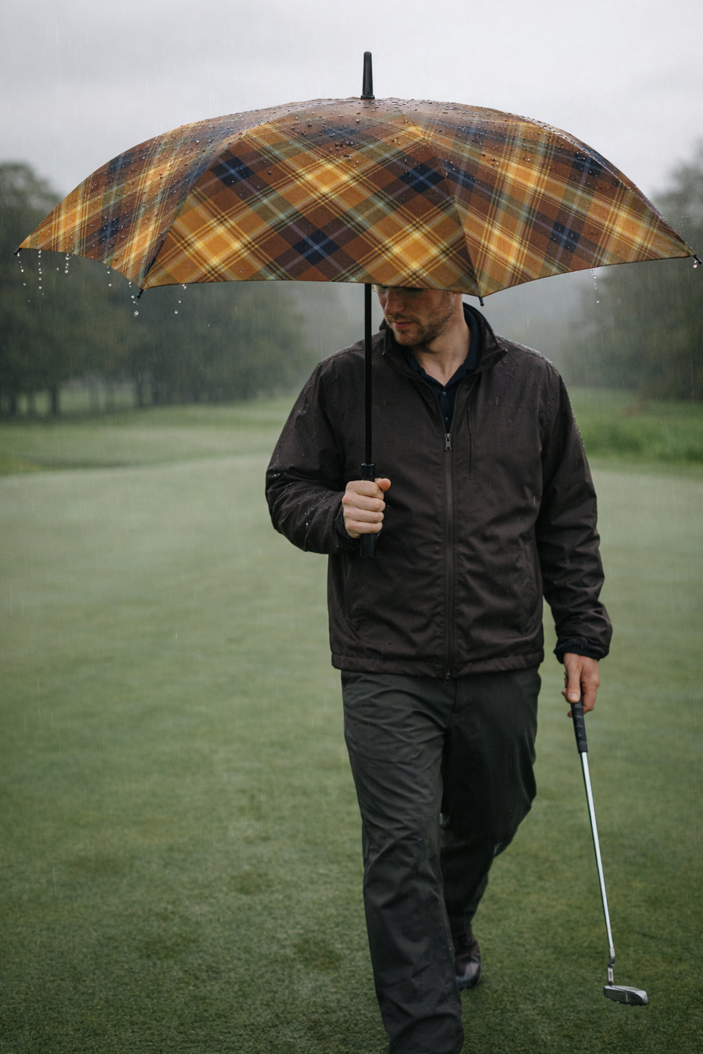 Golfer walking across wet fairway in steady rain beneath the Angel’s Share whisky-toned tartan umbrella, putter in hand and canopy beaded with droplets.